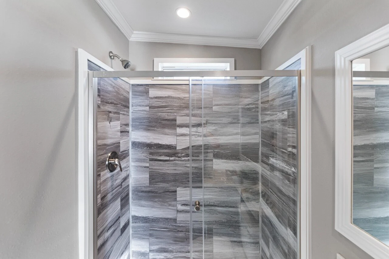 Modern shower with gray marble tile walls, glass doors, a chrome showerhead, and white crown molding.