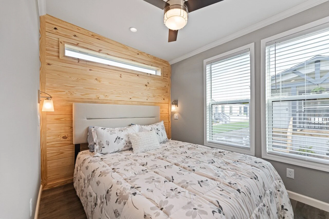 Modern bedroom with a wooden accent wall, bed with floral bedding, two windows with blinds, ceiling fan, and natural light.