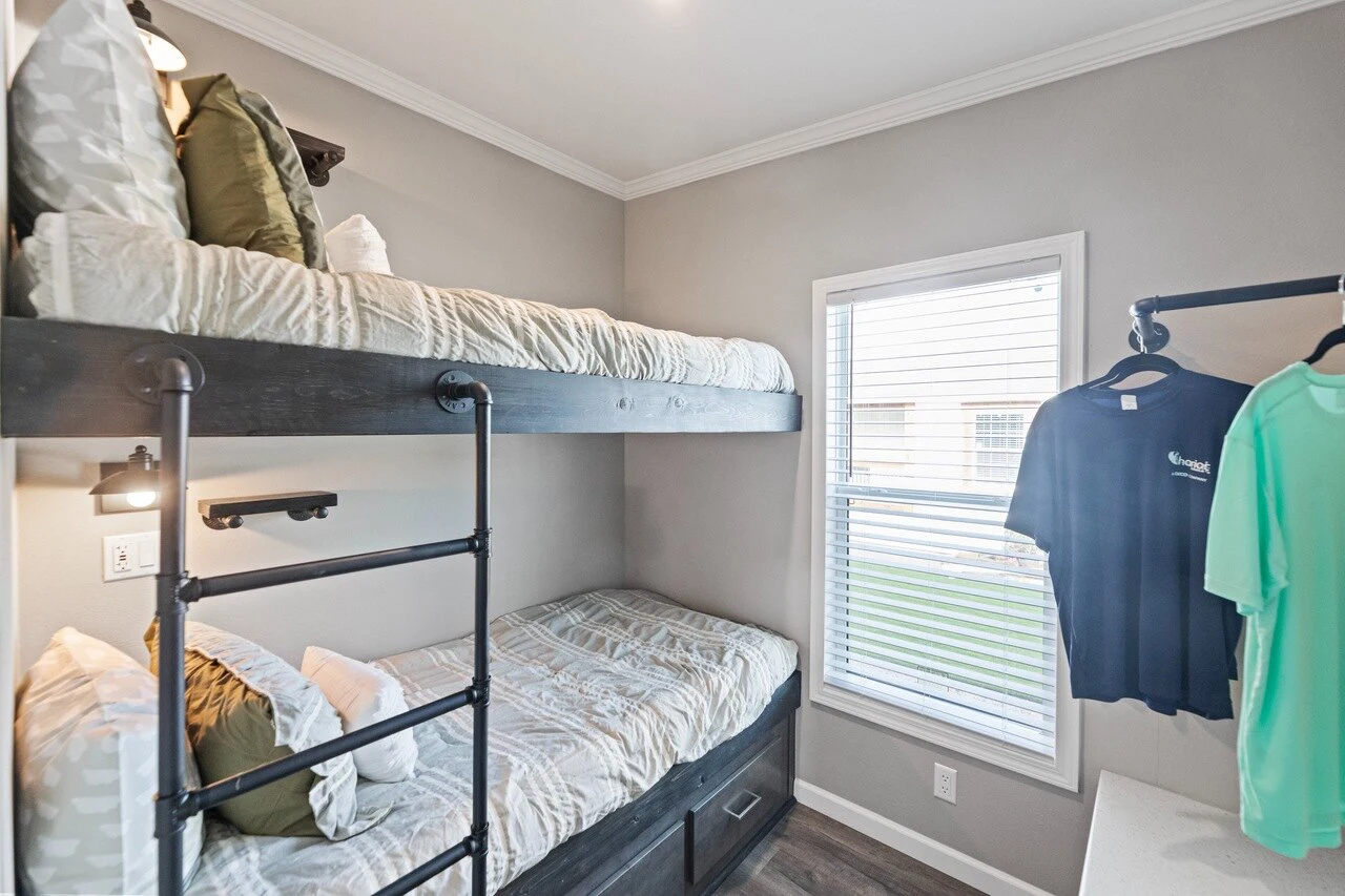 A small bedroom with dark wood bunk beds, patterned bedding, a window with blinds, a wall-mounted lamp, and two shirts hanging on a rack.