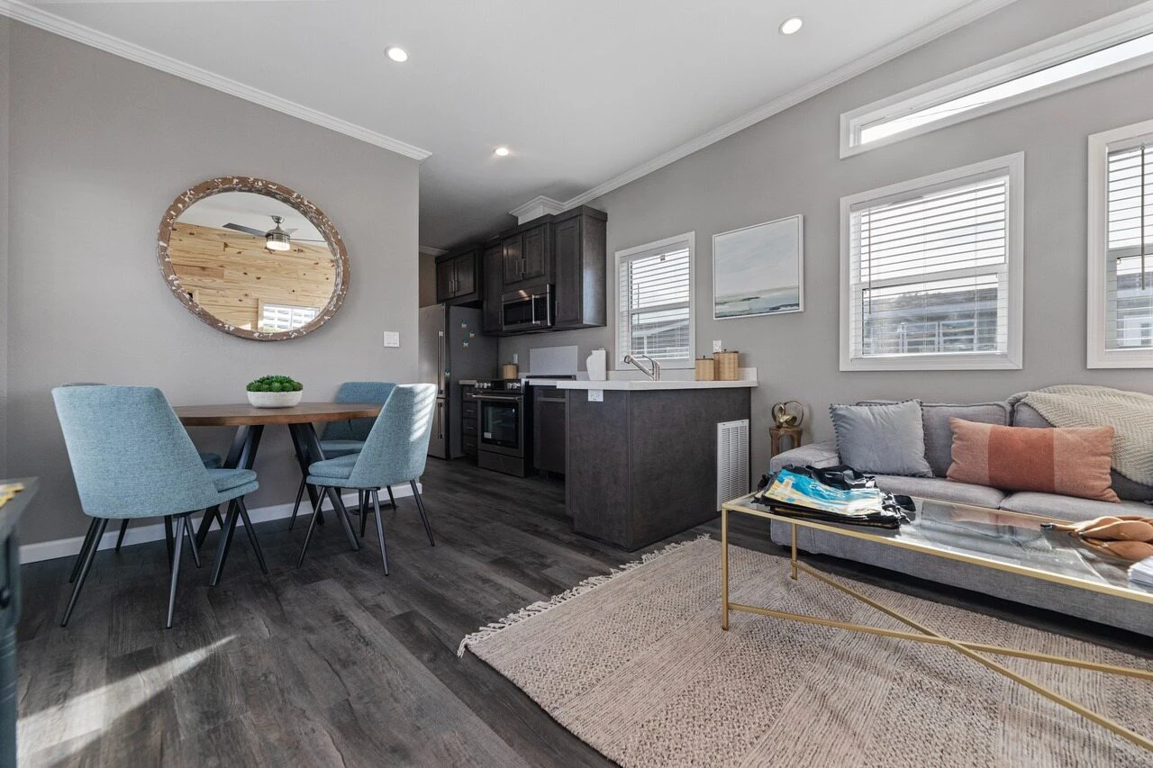 Modern open-plan living space with a small dining area, kitchen, and seating area featuring a couch, coffee table, and wall art. Room has gray walls, wood flooring, and natural light.