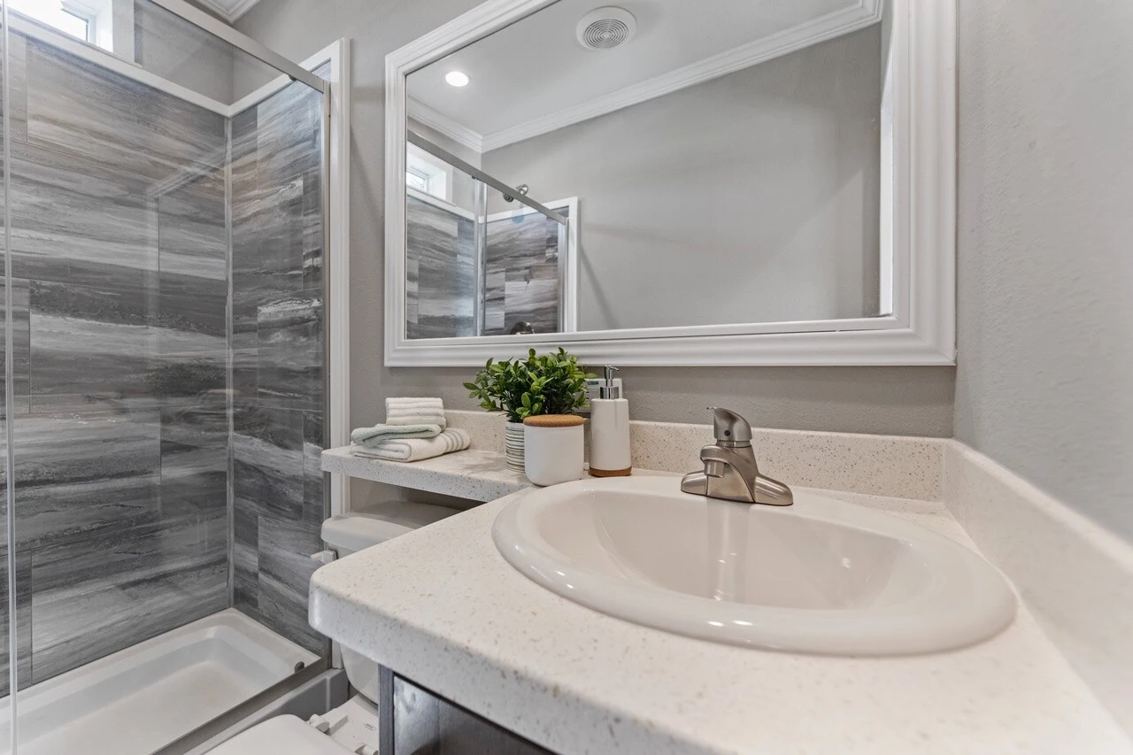 Modern bathroom with a glass shower, gray tile walls, white countertop with a sink, faucet, potted plant, and toiletries next to a large mirror.