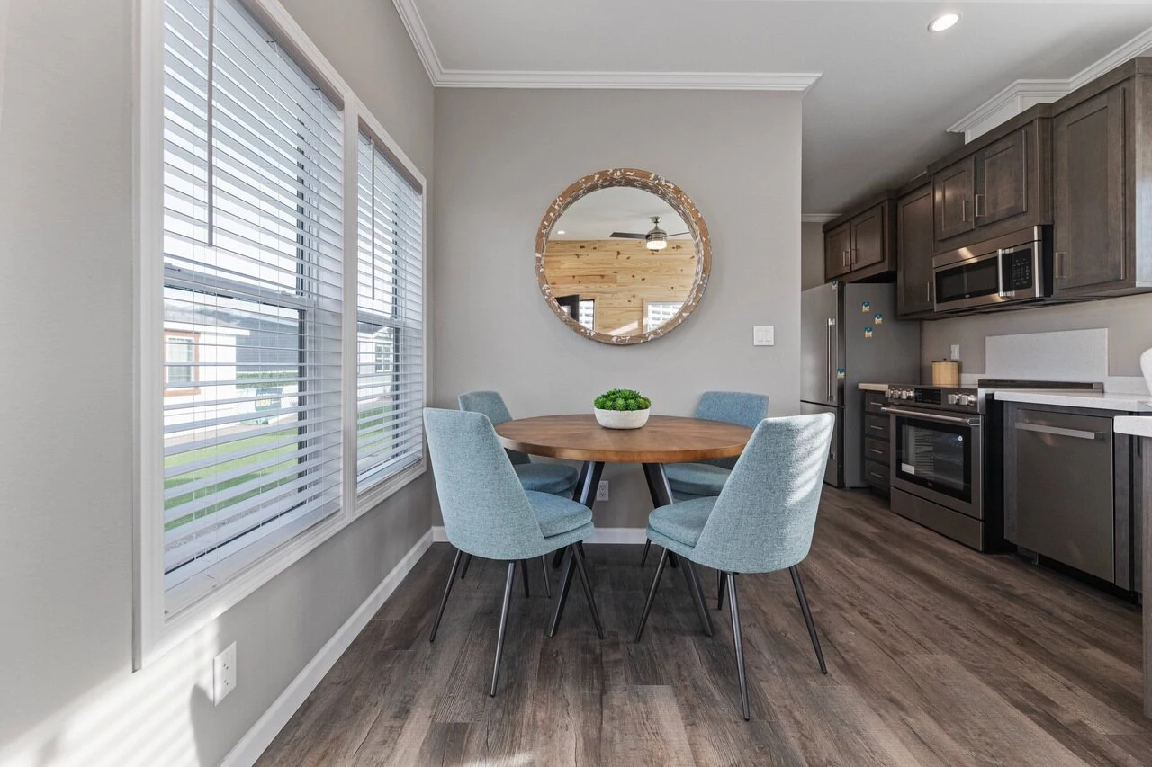 A modern kitchen with dark cabinets, stainless steel appliances, and a small round dining table with four blue chairs by a window. A round mirror hangs on the wall above the table.