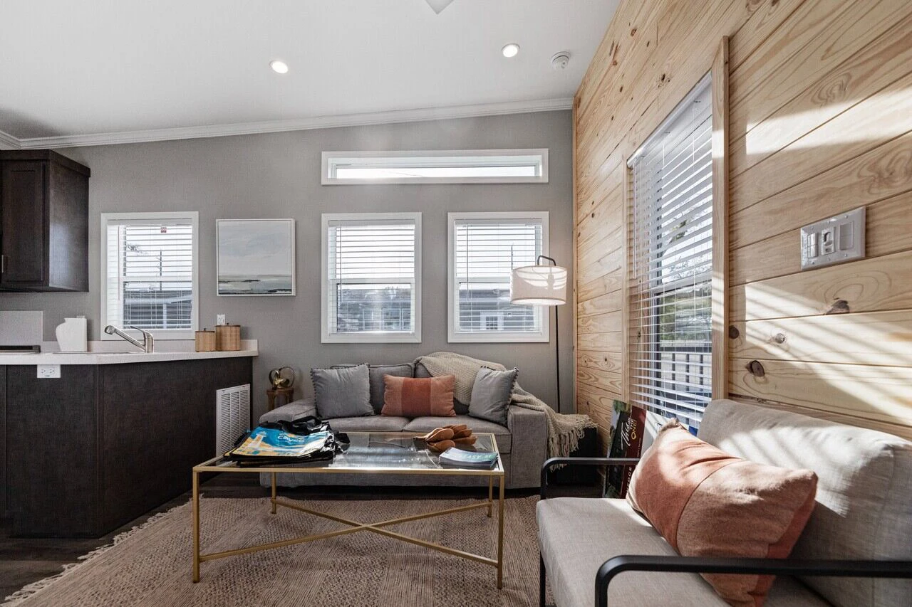 A modern living room with a gray sofa, a glass coffee table, a cushioned chair, and a wood-paneled accent wall, featuring large windows letting in natural light.