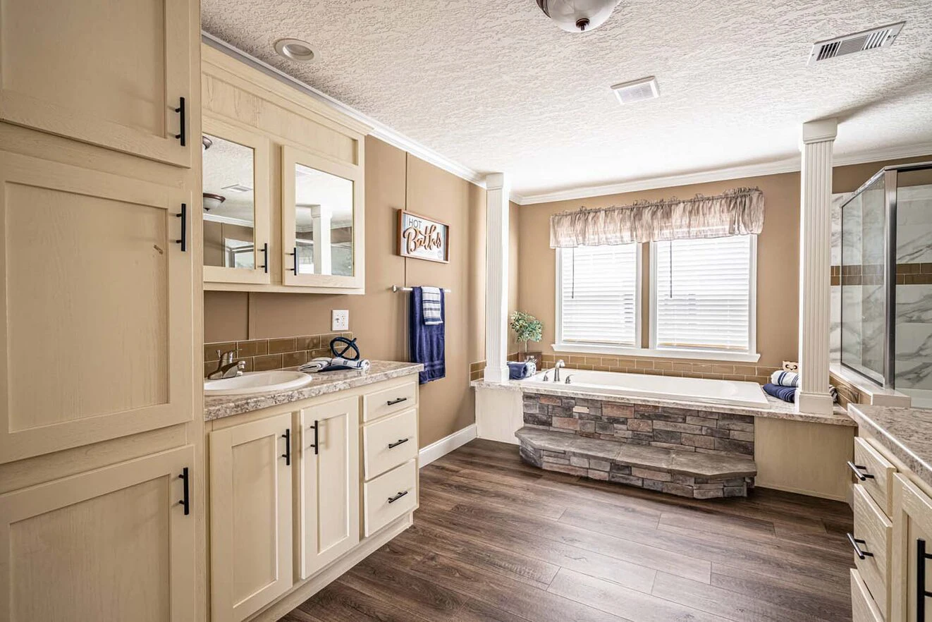 Spacious bathroom with light wood cabinets, a dual-sink vanity, large bathtub with stone surround, glass shower, and wood-look flooring. Natural light enters through two windows.