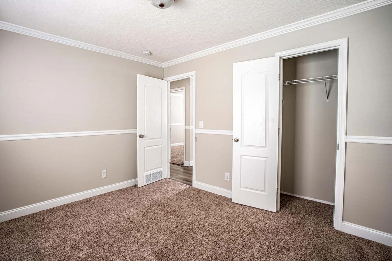 Empty beige-carpeted room with light taupe walls, white trim, two open white doors, and an empty closet with a shelf and hanging rod.