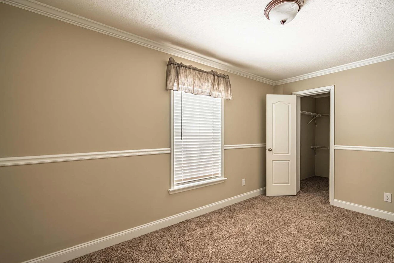 Empty beige-walled bedroom with a window, white blinds, carpeted floor, ceiling light, and an open door leading to a small walk-in closet.
