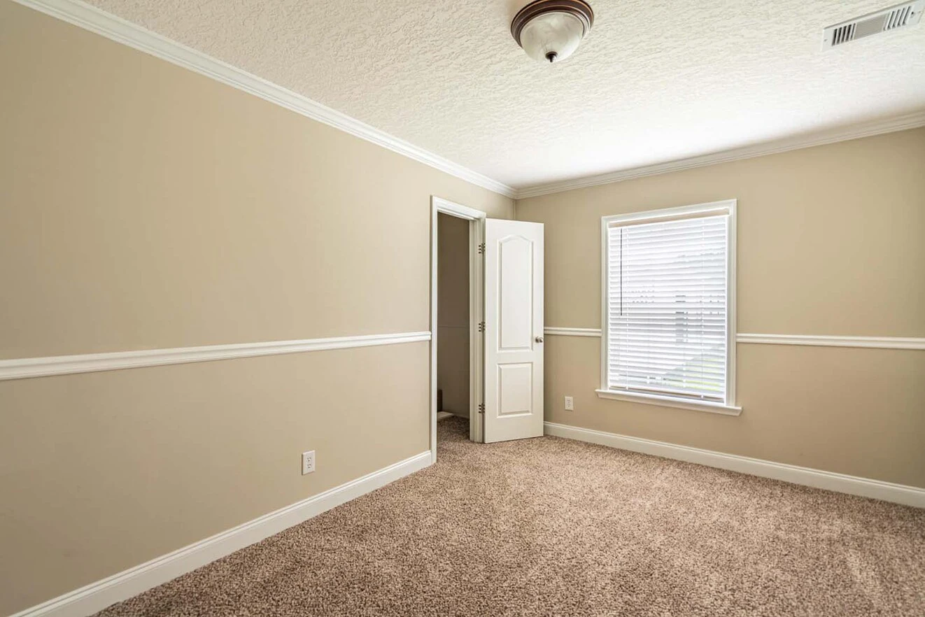 Empty beige room with white trim, carpet flooring, a closed window with blinds, and an open door leading to another area. Ceiling light fixture is visible.