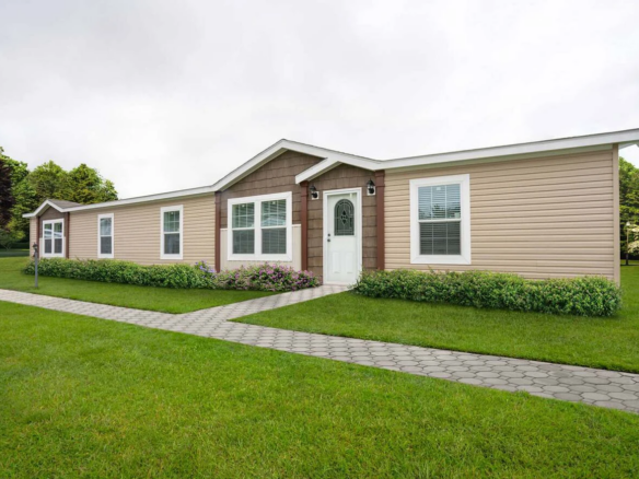 A single-story manufactured home with tan and brown siding, white trim, central front door, multiple windows, and a paved walkway, surrounded by green lawn and shrubs.