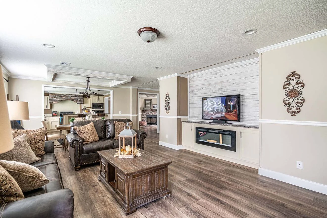 Bright living room with brown leather sofas, wooden coffee table, mounted TV, electric fireplace, and decorative wall accents; kitchen visible in the background.