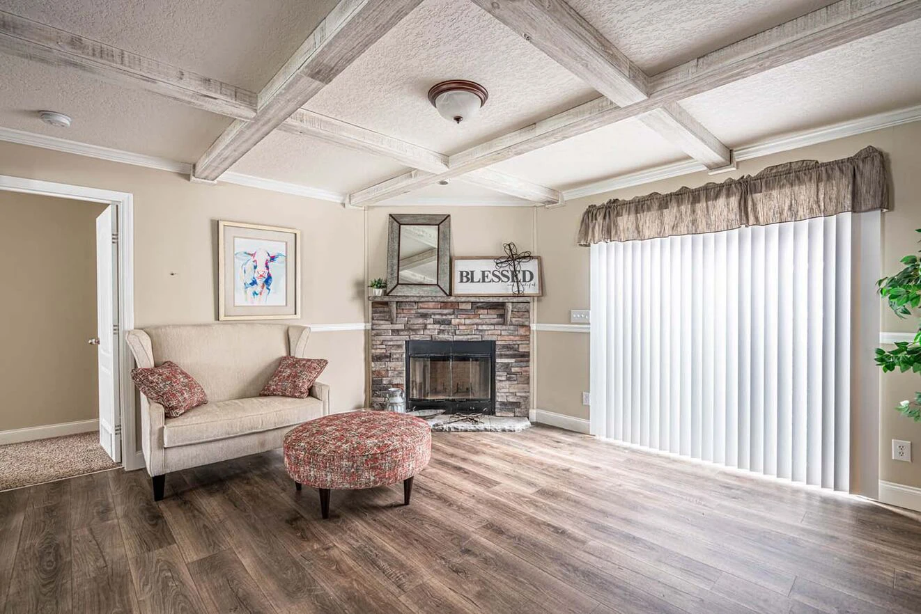 A living room with wood flooring, a beige armchair, a floral ottoman, a stone fireplace, a "Blessed" sign, wall art, and large vertical blinds covering a window.