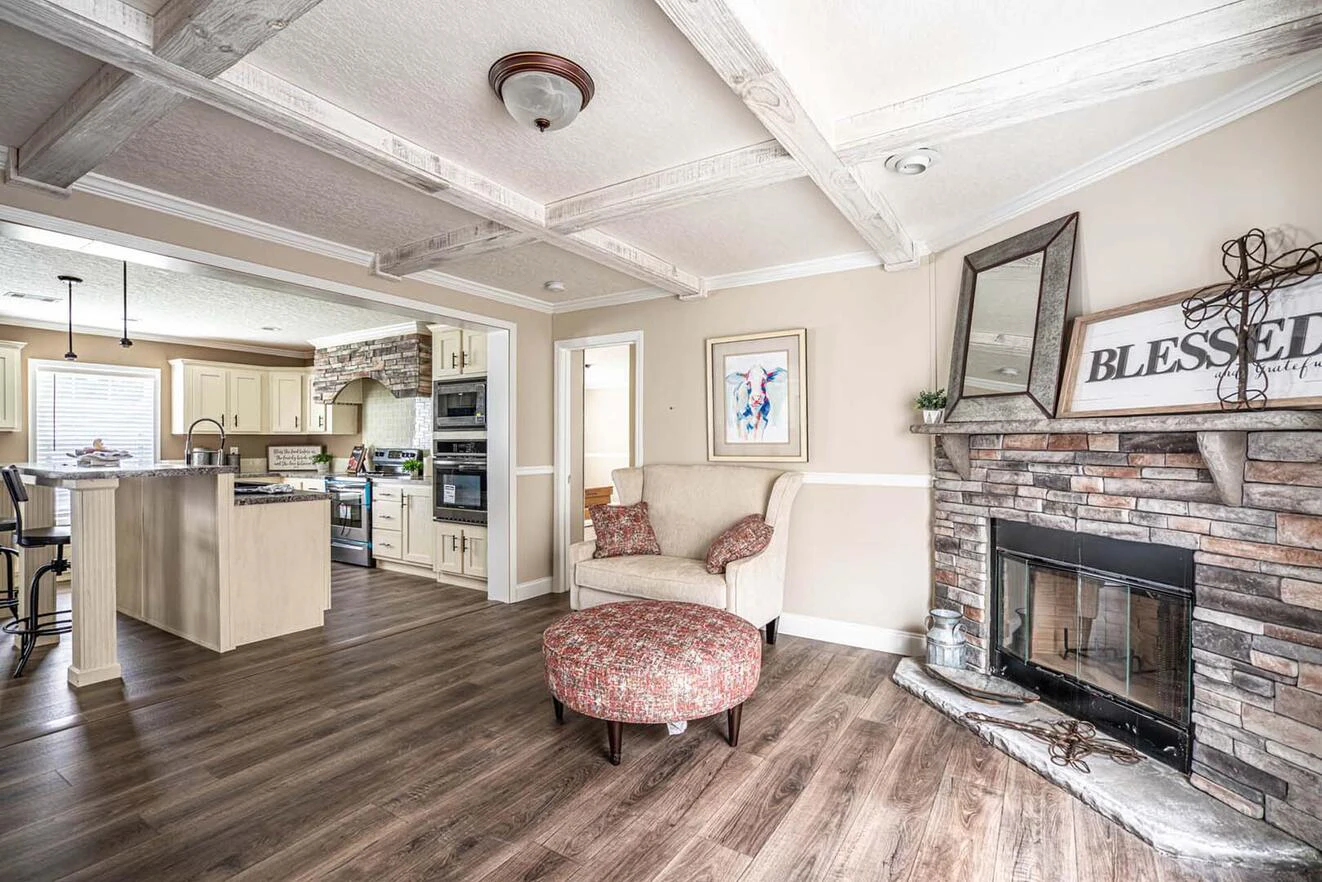 A cozy living room with wood floors, a stone fireplace, an armchair with ottoman, and an open view of a kitchen with cream cabinets and stainless steel appliances.