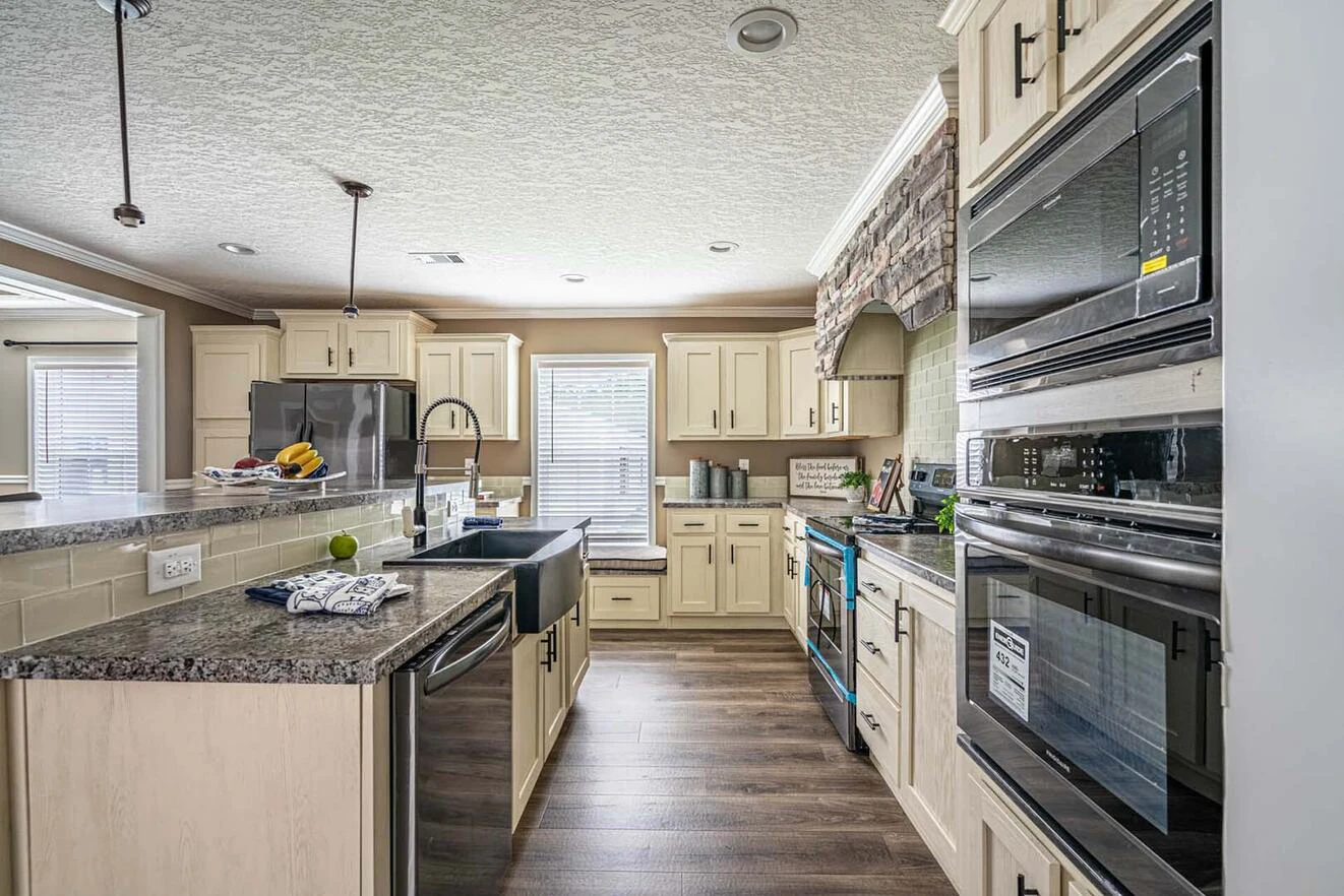 Modern kitchen with cream cabinets, granite countertops, stainless steel appliances, a farmhouse sink, double ovens, and wood flooring. Natural light enters through two windows.