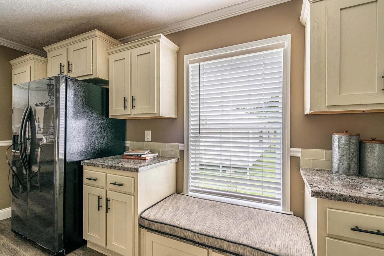 A kitchen with cream-colored cabinets, a black refrigerator, granite countertops, and a cushioned window seat below a window with closed blinds.