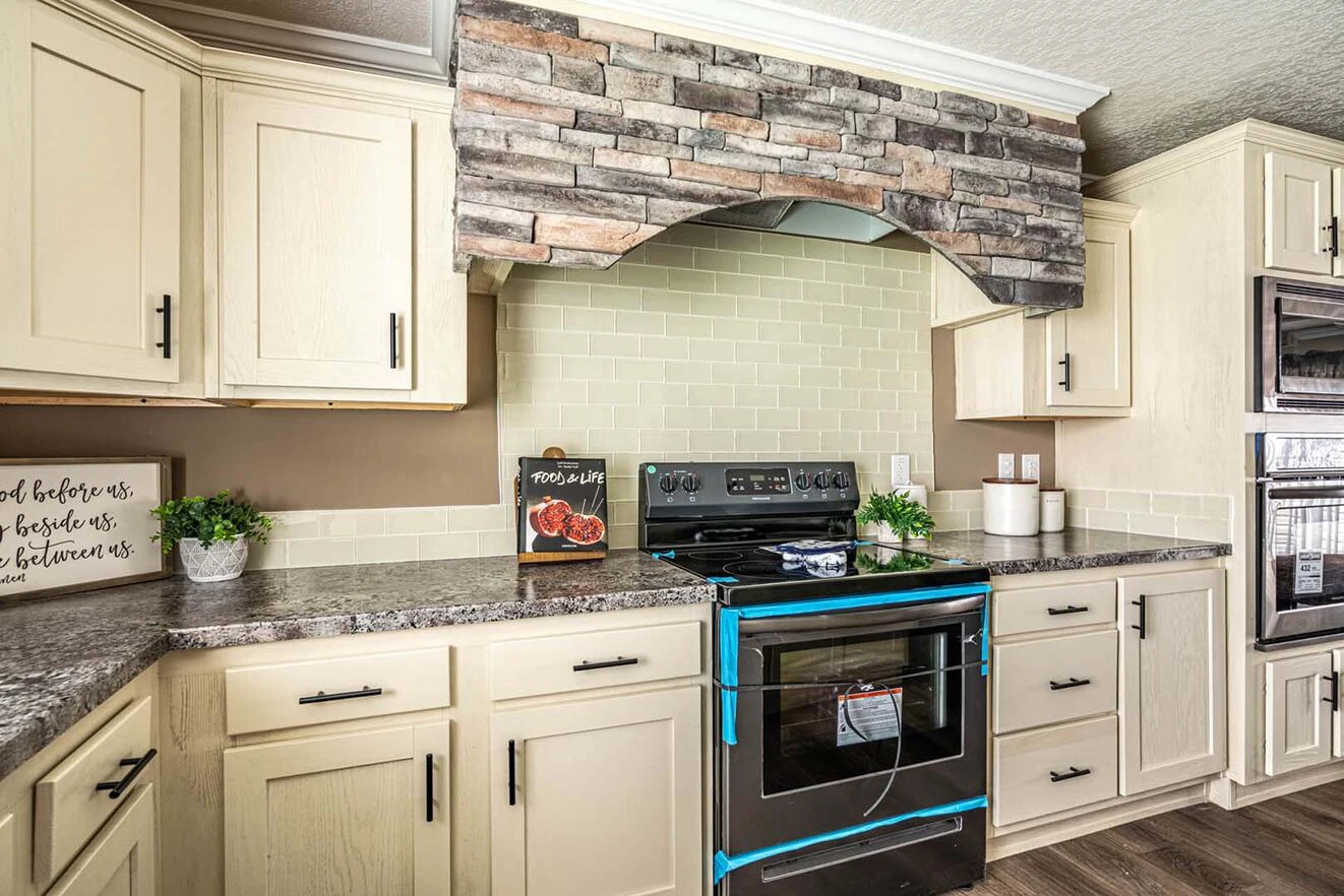 A modern kitchen features light wood cabinets, a stone range hood, a stove with protective blue tape, beige tile backsplash, and decorative items on the countertop.