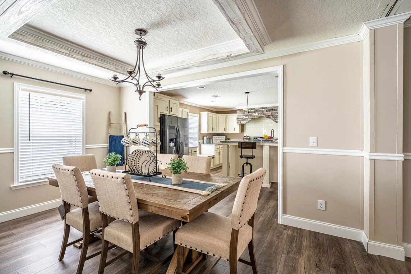 A dining area with a wooden table, six upholstered chairs, decorative centerpiece, and an open view into a kitchen with light cabinets and stainless steel appliances.