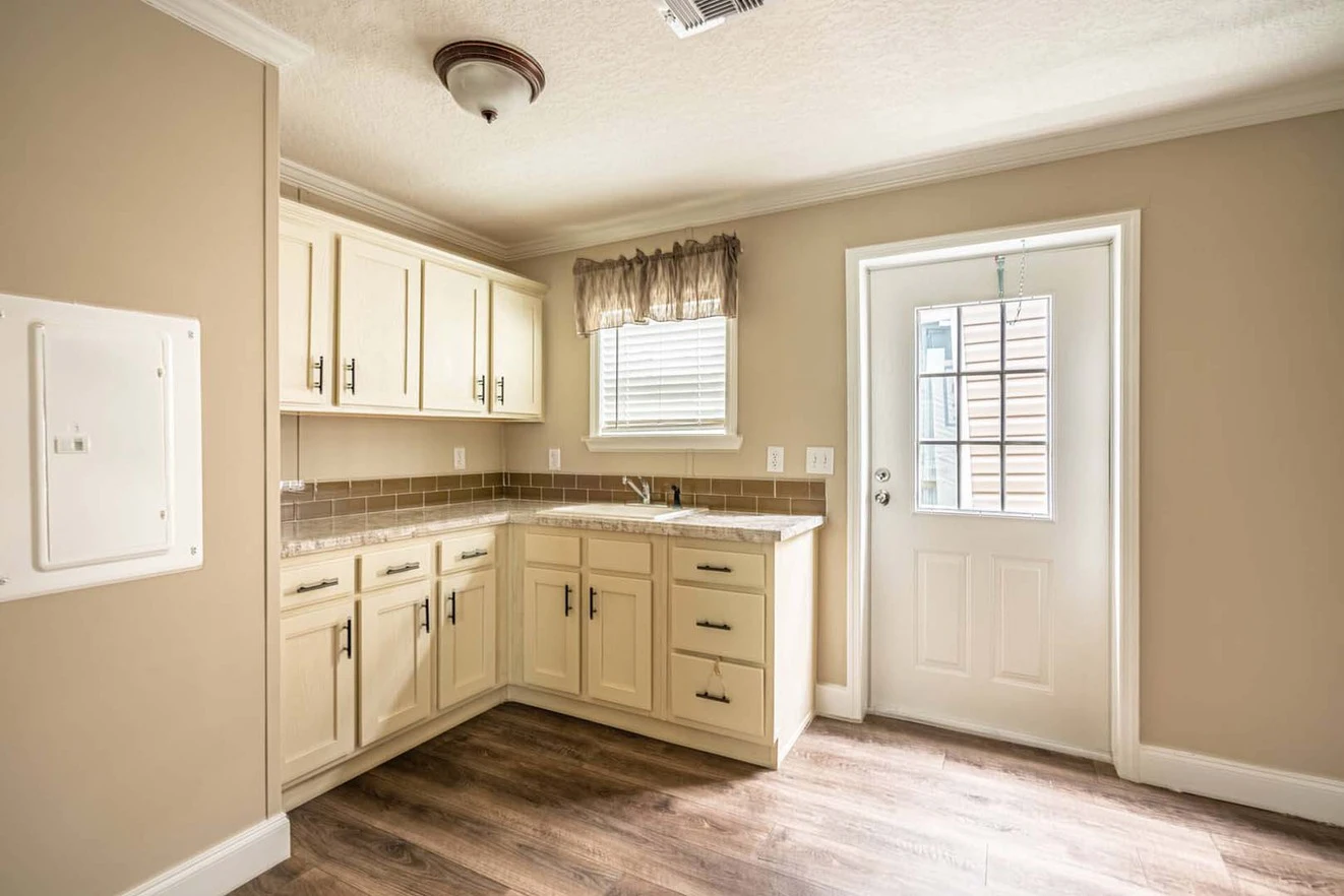 Small kitchen with cream cabinets, brown tile backsplash, single sink under a window, and a door leading outside. The floor is wood-patterned laminate.