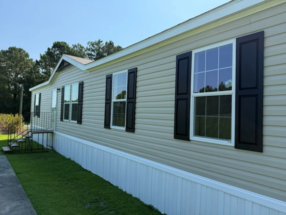 A beige manufactured home with black shutters and white skirting, set on a grassy lawn with trees in the background.