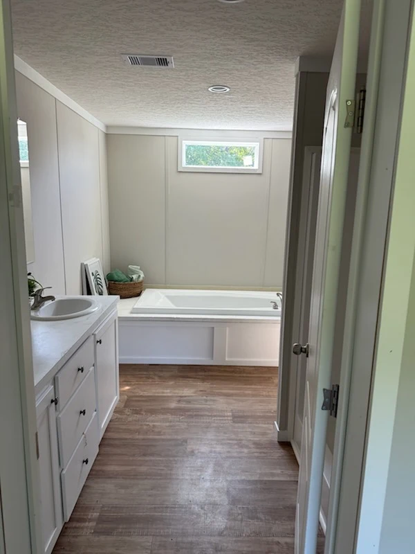A bathroom with wood flooring, a white vanity with sink, and a built-in bathtub beneath a small window.