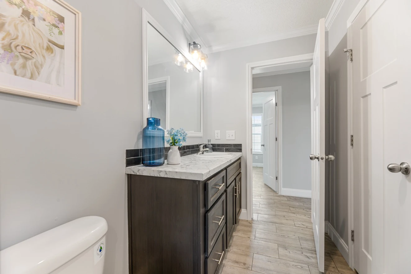 Modern bathroom with a dark wood vanity, marble countertop, wall mirror, blue glass vase, and light gray walls; hallway visible through open door.