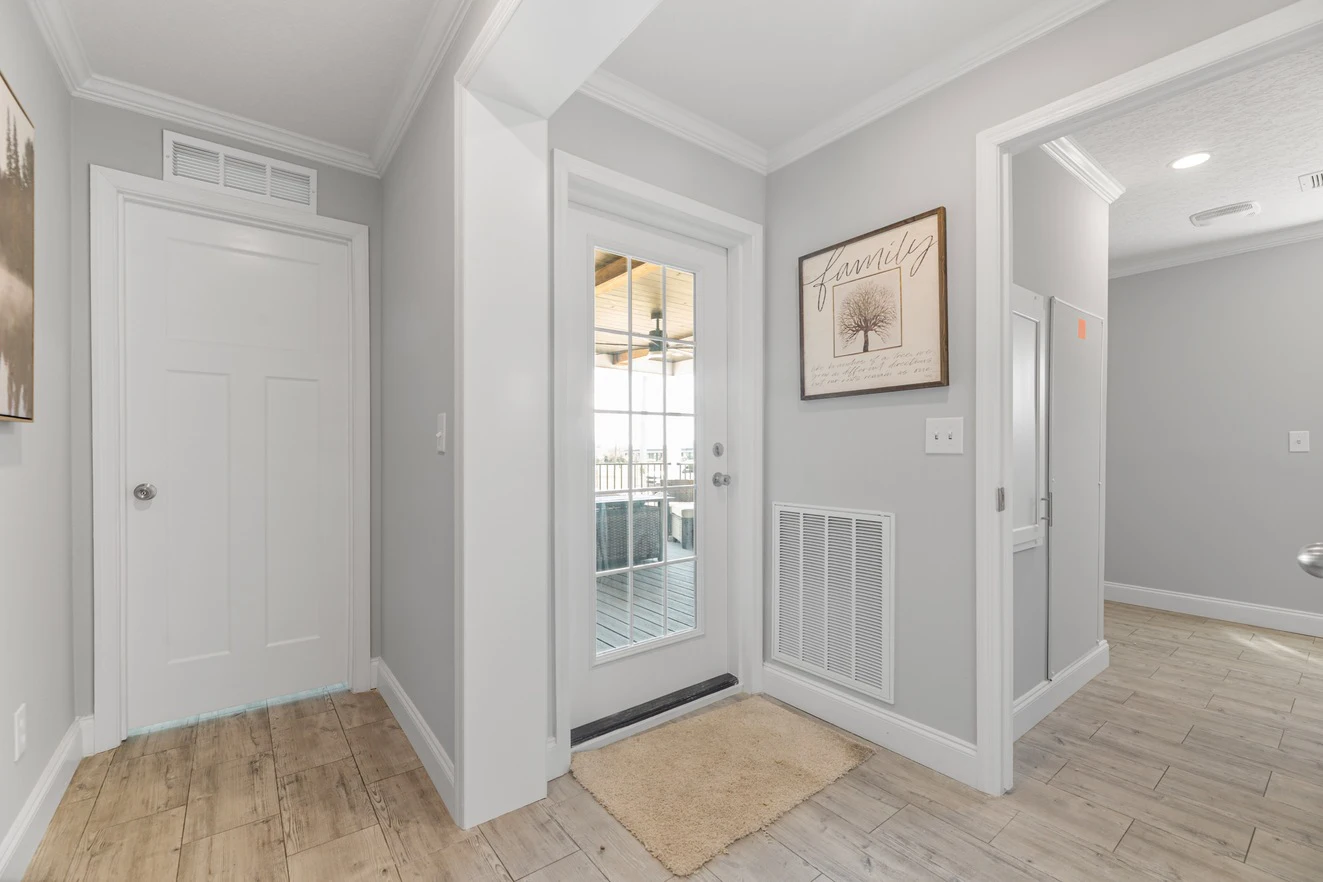 Bright hallway with light wood flooring, white walls, a door leading outside, framed wall art, and a beige mat in front of the door.
