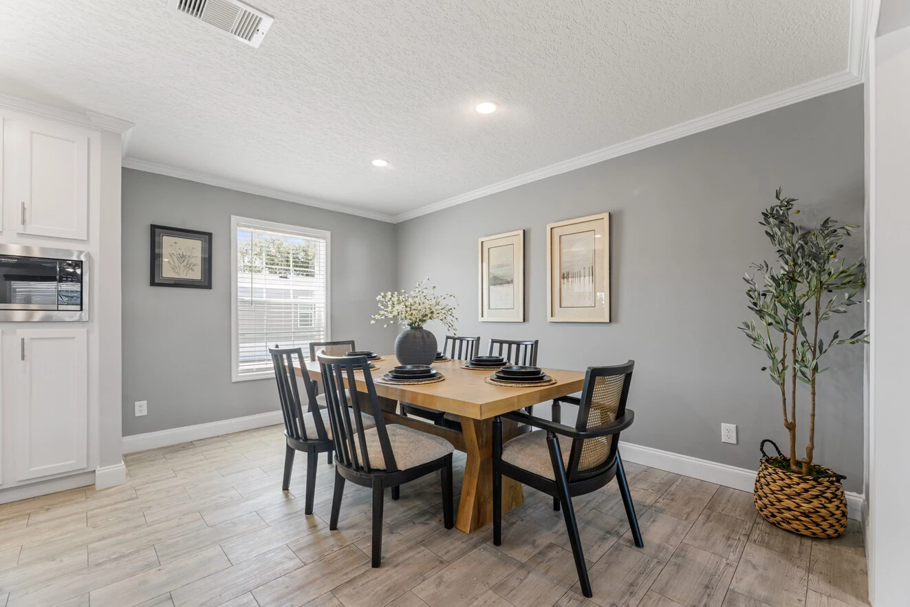Modern dining room with a wooden table, six black chairs, minimalist decor, potted plant, and framed artwork on gray walls, illuminated by natural light from a window.