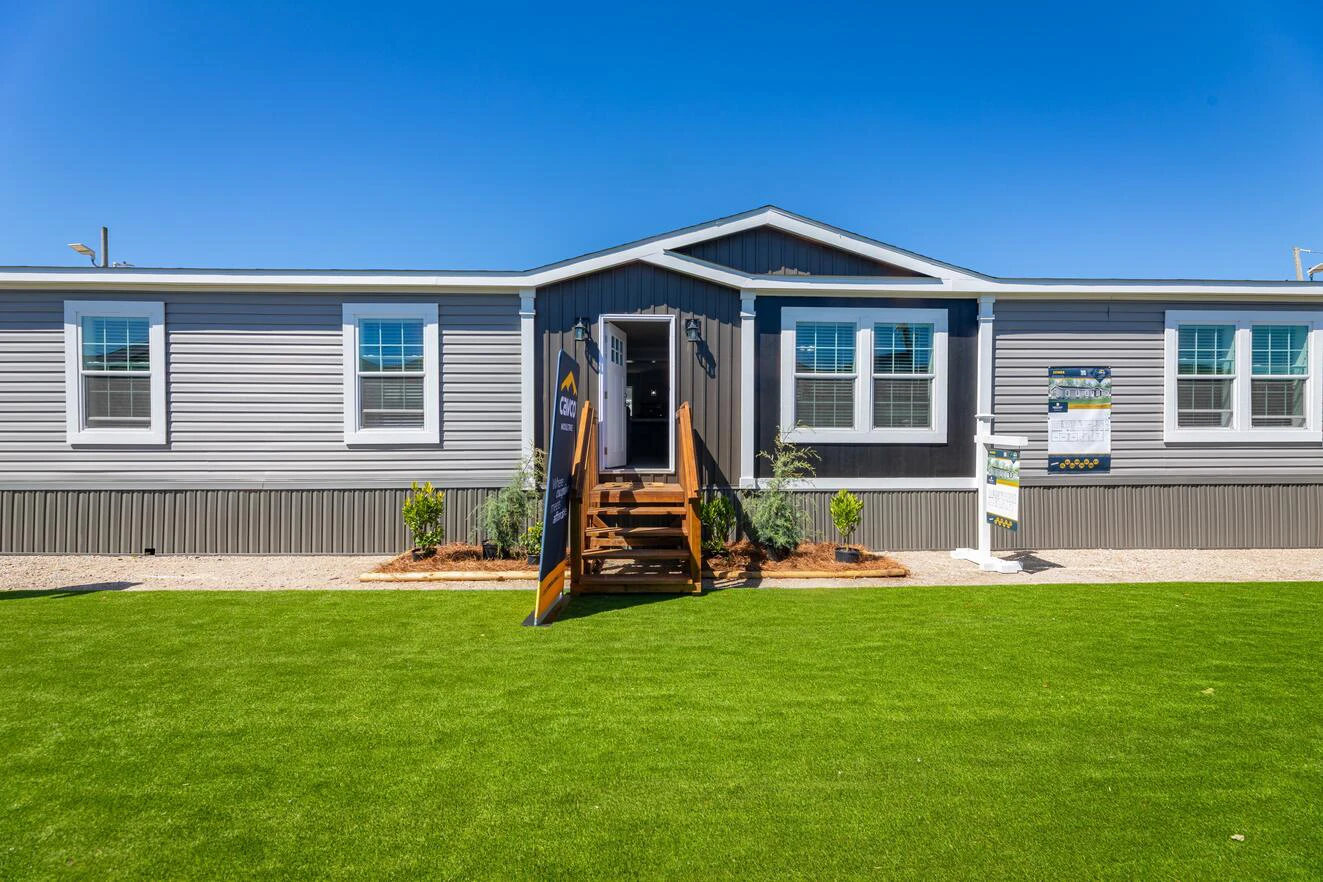 A modern manufactured home with gray siding, white trim, and a central entryway sits on a lawn under a clear blue sky.