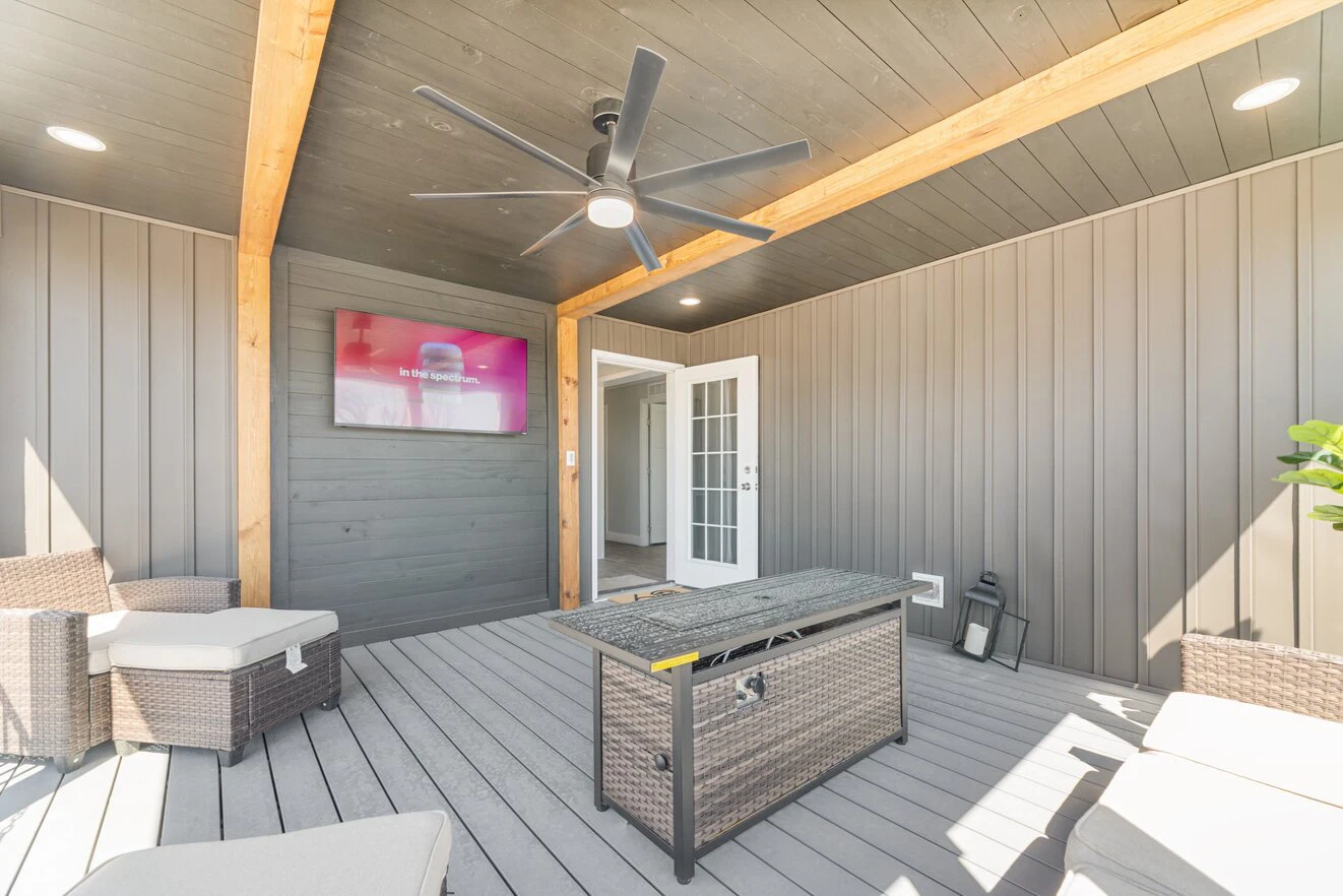 A covered patio with wicker lounge furniture, a ceiling fan, a TV mounted on the wall, and a glass door leading inside.