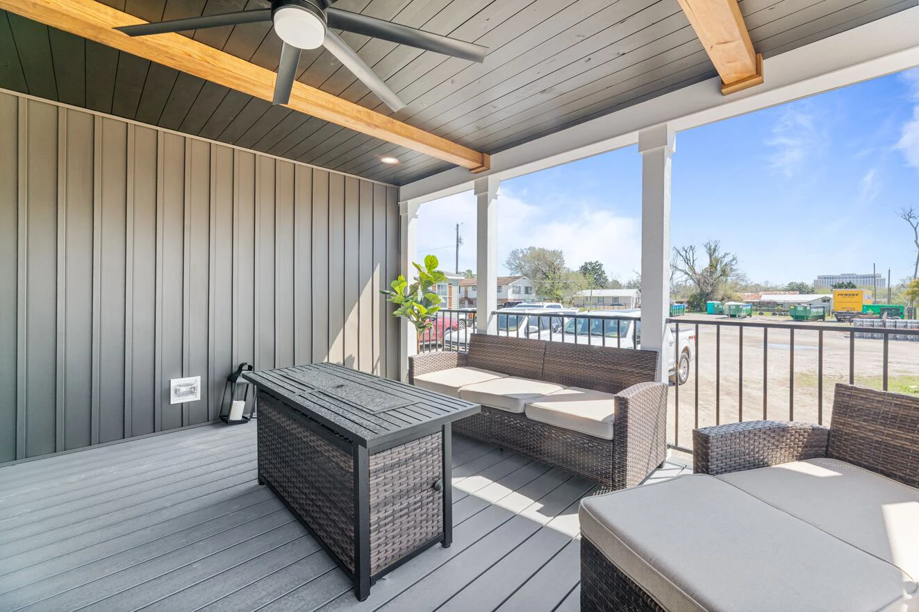 Covered outdoor patio with rattan furniture, a ceiling fan, wood beams, a fire table, and a view of a parking lot and buildings in the background.
