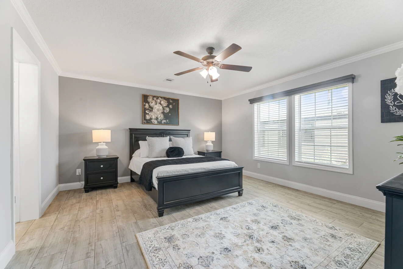 A bedroom with a queen bed, two nightstands with lamps, a ceiling fan, large windows with blinds, and a patterned rug on a wooden floor.