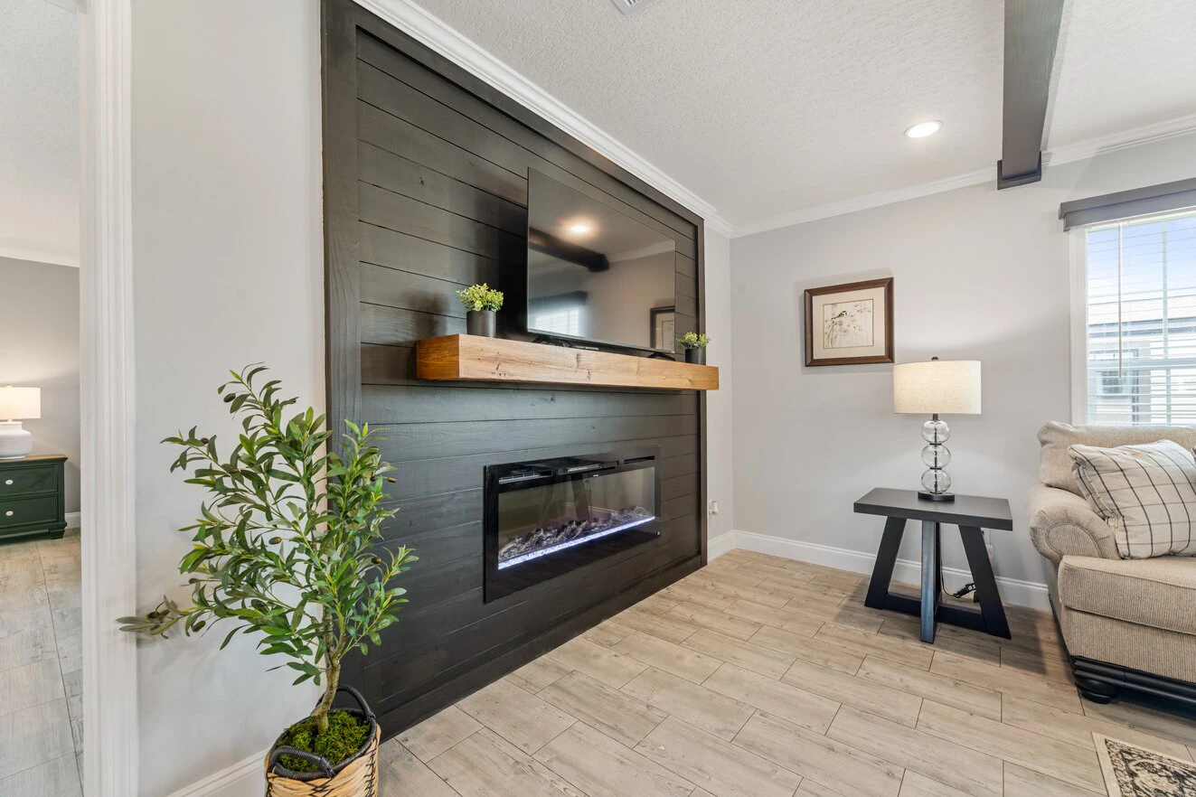 Modern living room with a black accent wall, mounted TV, wooden mantel, electric fireplace, potted plant, lamp, and beige sofa.