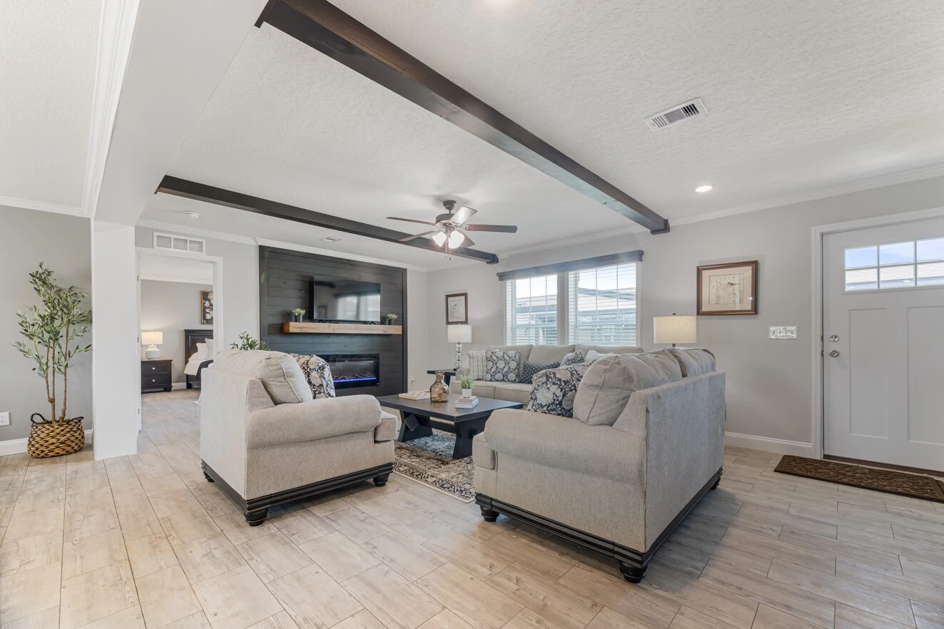 Bright living room with two beige sofas, a coffee table, wood floor, exposed ceiling beams, fireplace, and large windows. An open doorway leads to another room.