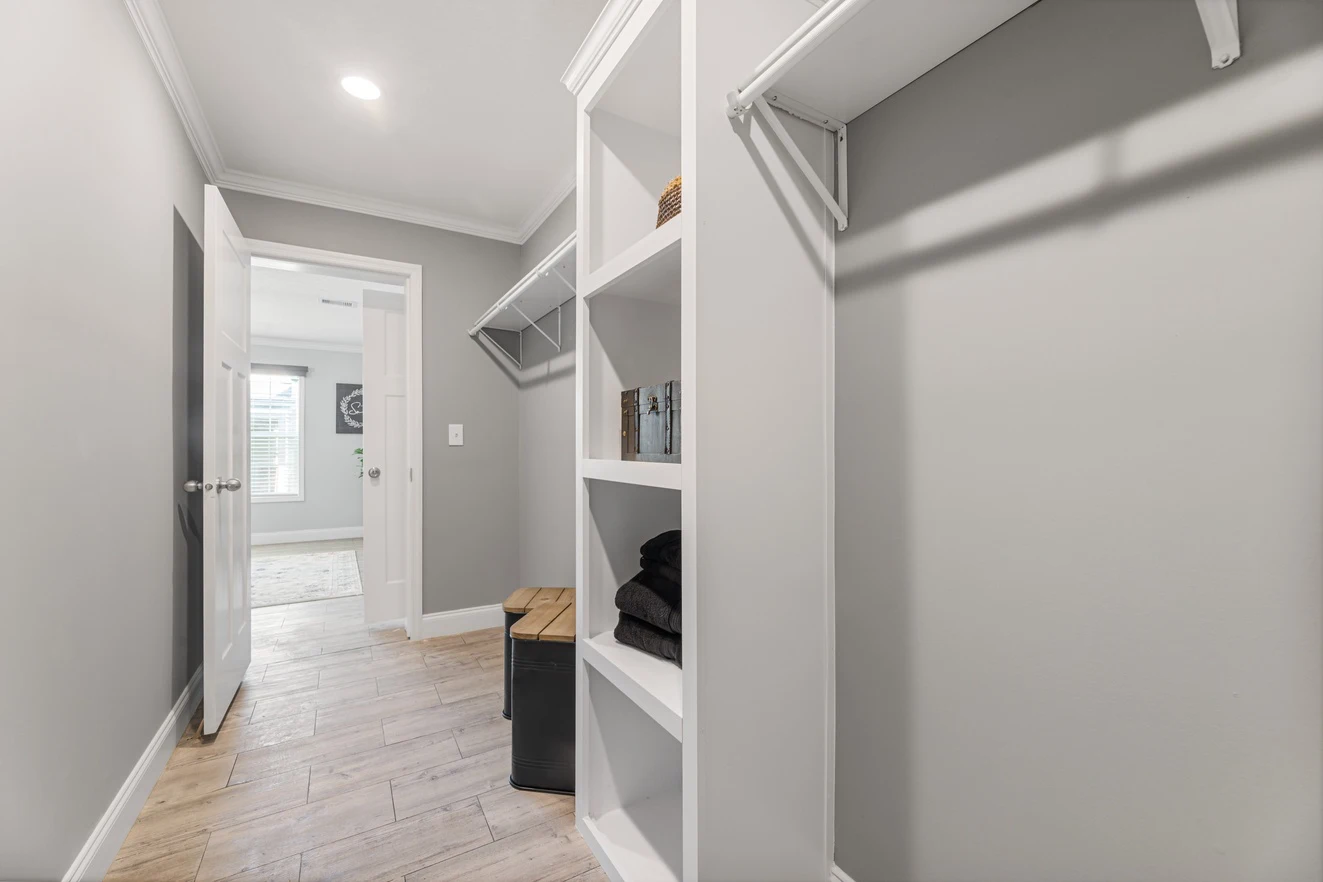 A modern walk-in closet with built-in white shelves, hanging rods, gray walls, wood-look floor, and a bench; door open to an adjoining room with natural light.