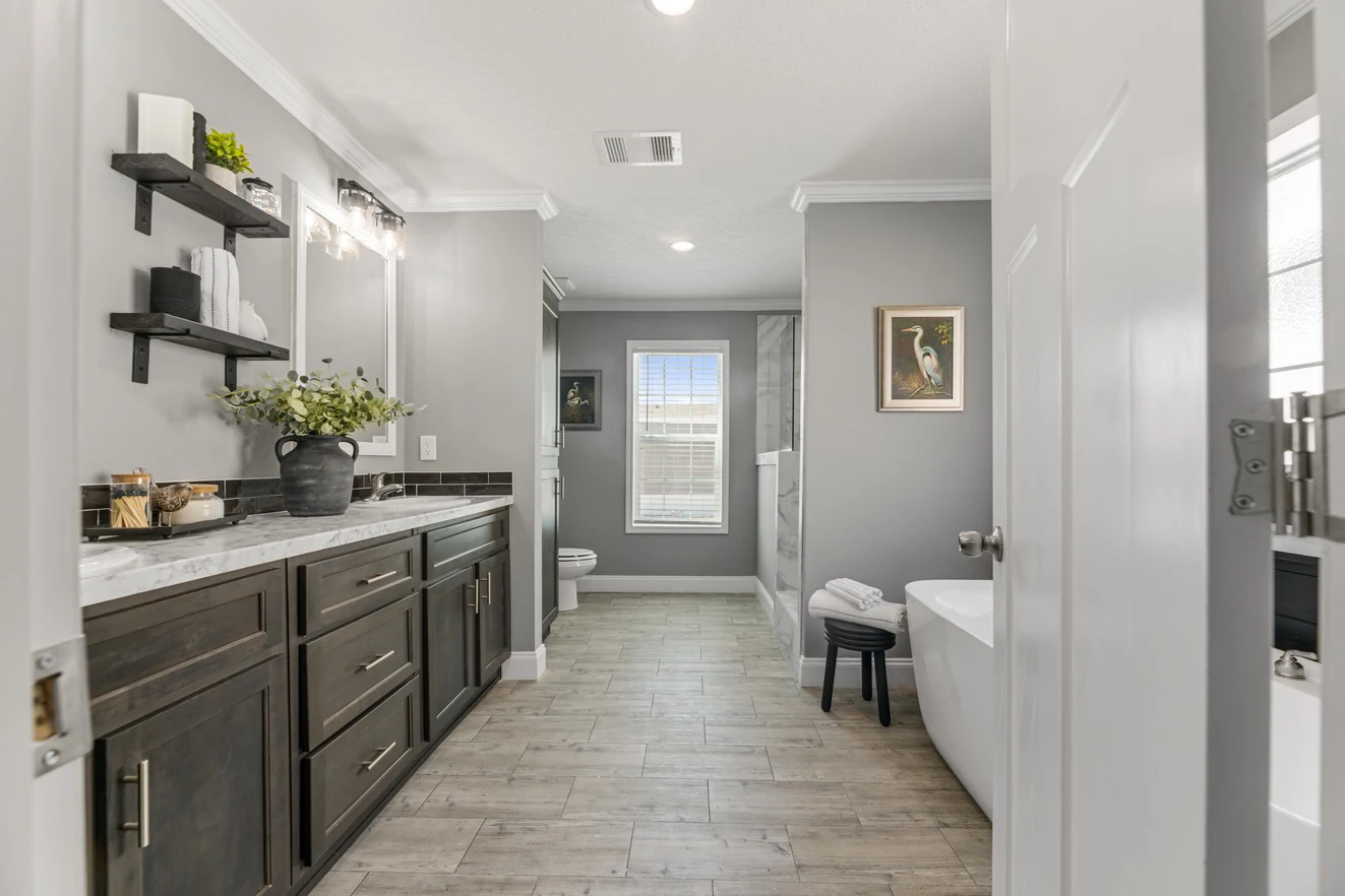 Modern bathroom with gray walls, double sink vanity, freestanding bathtub, shelves with decor, and a window at the far end letting in natural light.