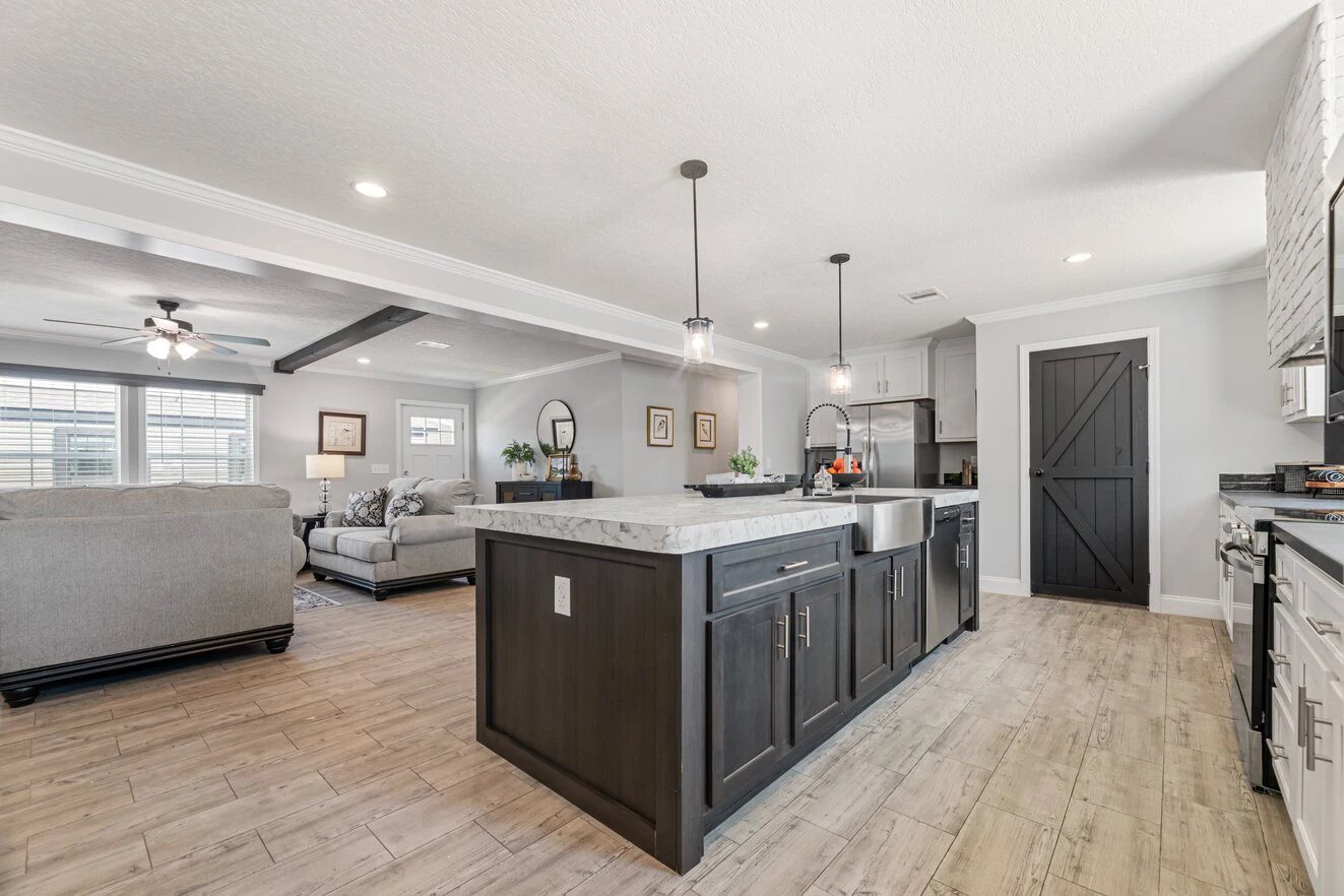 Modern kitchen with a dark island, farmhouse sink, pendant lighting, and adjacent living room featuring light gray sofas and wood flooring.