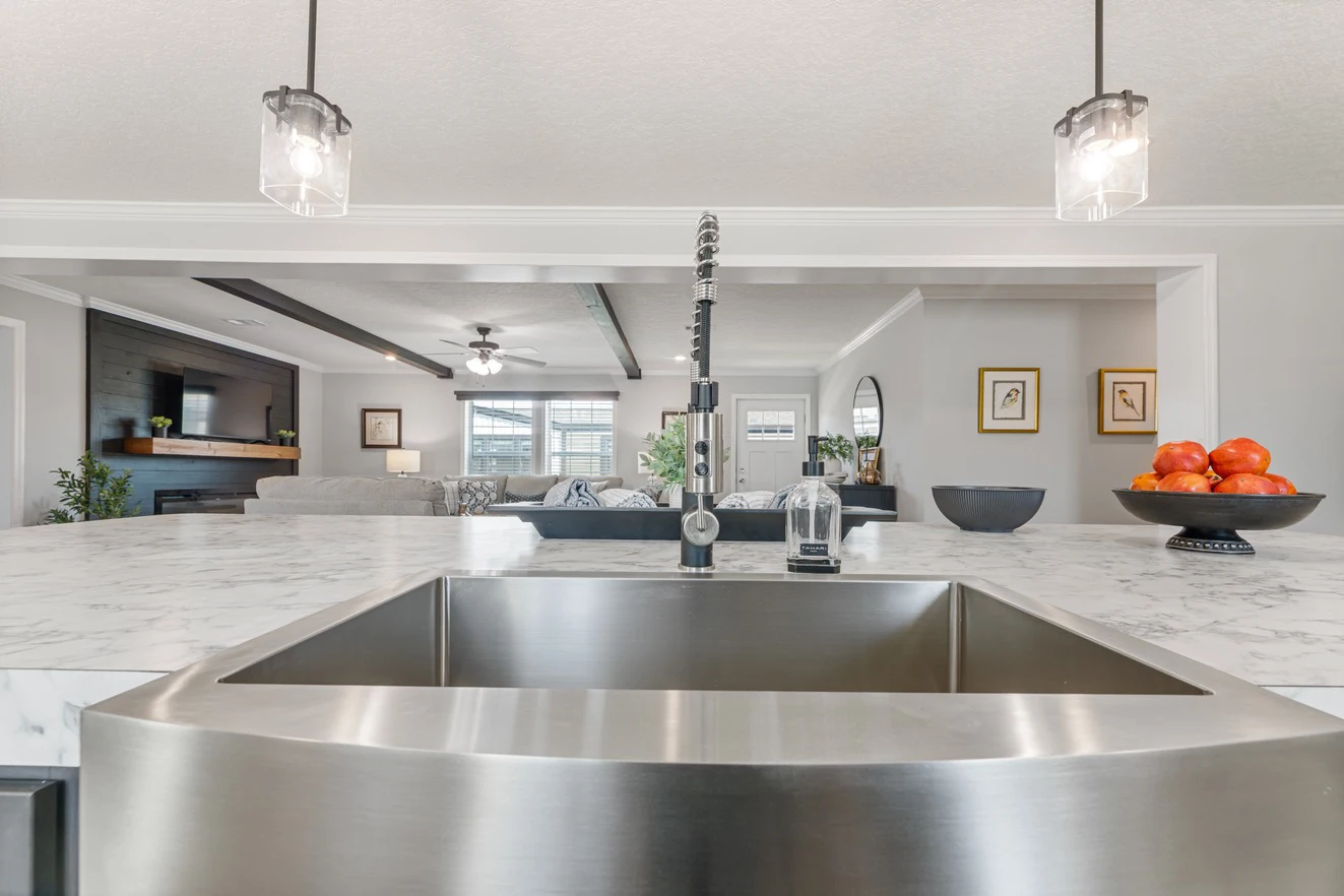 Modern kitchen with a marble island, stainless steel sink, fruit bowl, and pendant lights, overlooking an open living room with a TV and ceiling fan.