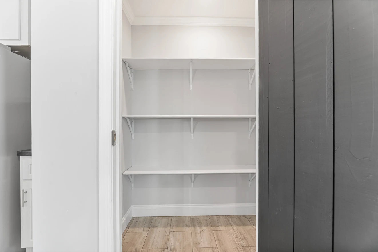 Empty walk-in pantry with three white shelves, white walls, light wood flooring, and partial view of adjacent dark and white cabinetry.