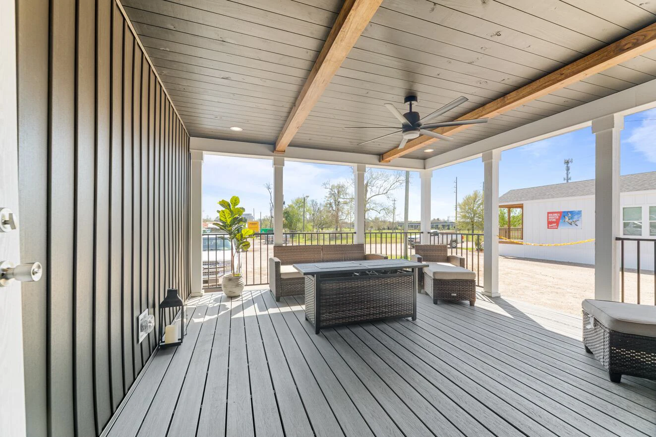 Covered porch with gray decking, outdoor wicker furniture, potted plants, black metal railing, ceiling fan, and view of a street with a small building outside.
