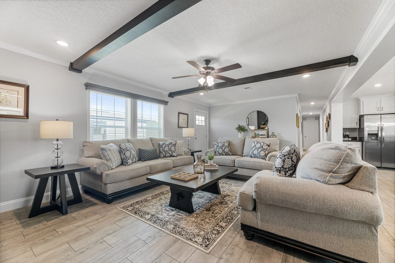 Spacious living room with beige sofas, patterned rug, coffee table, ceiling fan, exposed beams, large windows, and light wood flooring. Kitchen visible in the background.