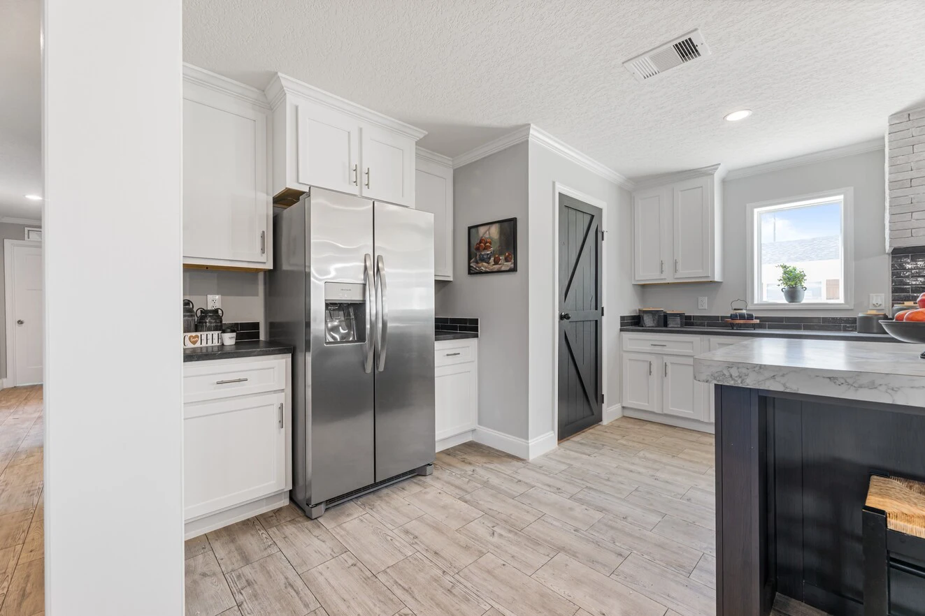 Modern kitchen with stainless steel refrigerator, white cabinets, black accents, wood-style tile flooring, and a gray barn-style pantry door.