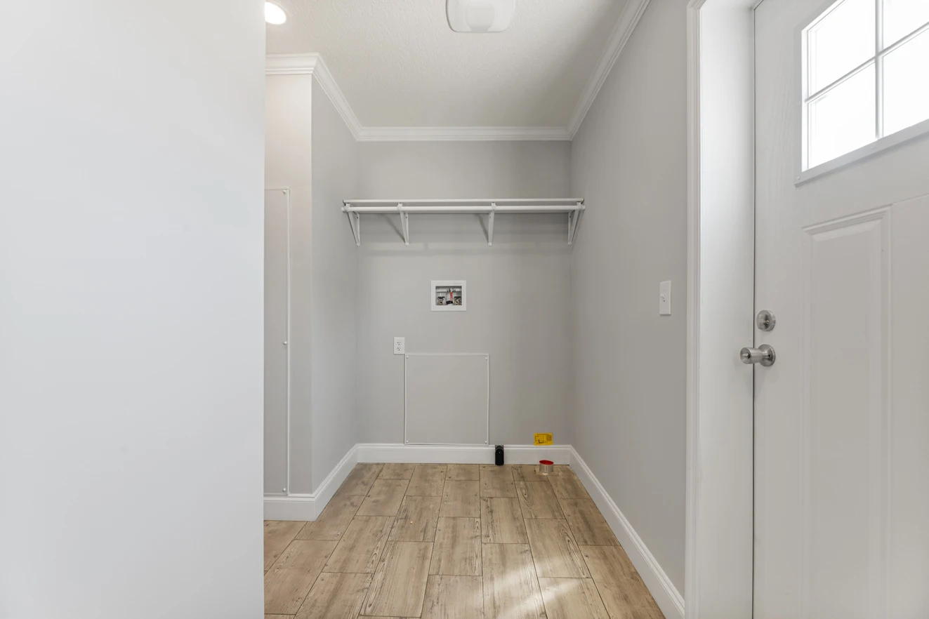 Empty laundry room with light gray walls, a wooden floor, a white shelf, and hookups for a washer and dryer; a white door is on the right side.