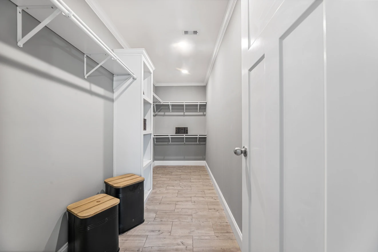 Empty walk-in closet with white shelves, hanging rods, and two black bins with wooden lids on a light wood floor.