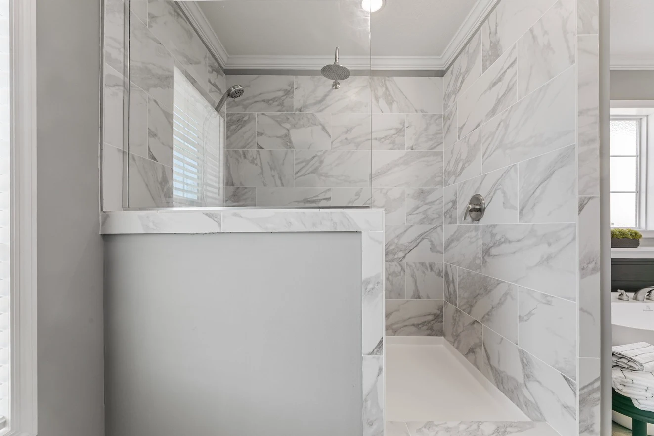 Modern walk-in shower with white and gray marble tiles, glass divider, chrome showerhead, and a window providing natural light.