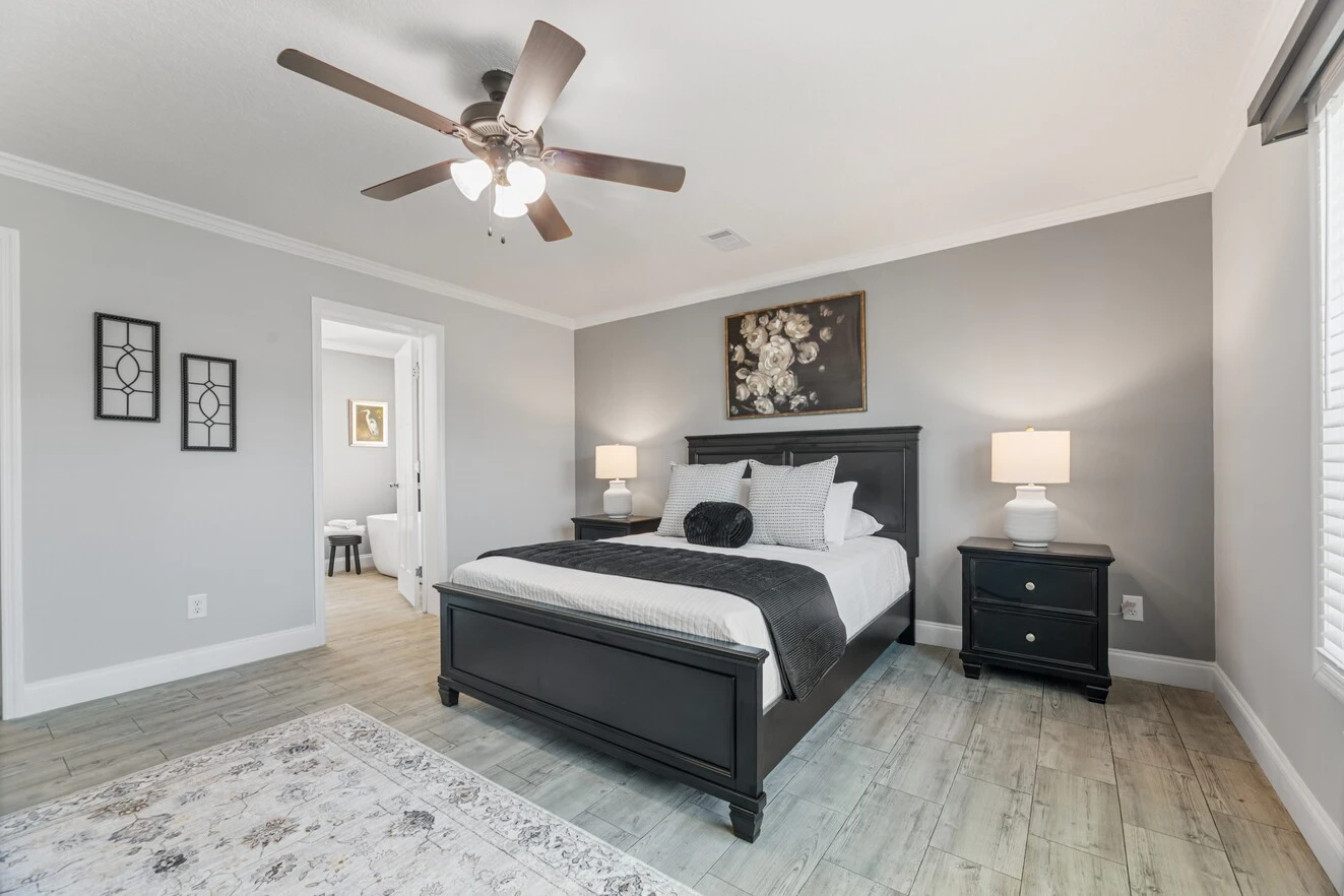 A modern bedroom with a dark wood bed frame, two matching nightstands with lamps, a ceiling fan, and neutral decor in gray and white tones.