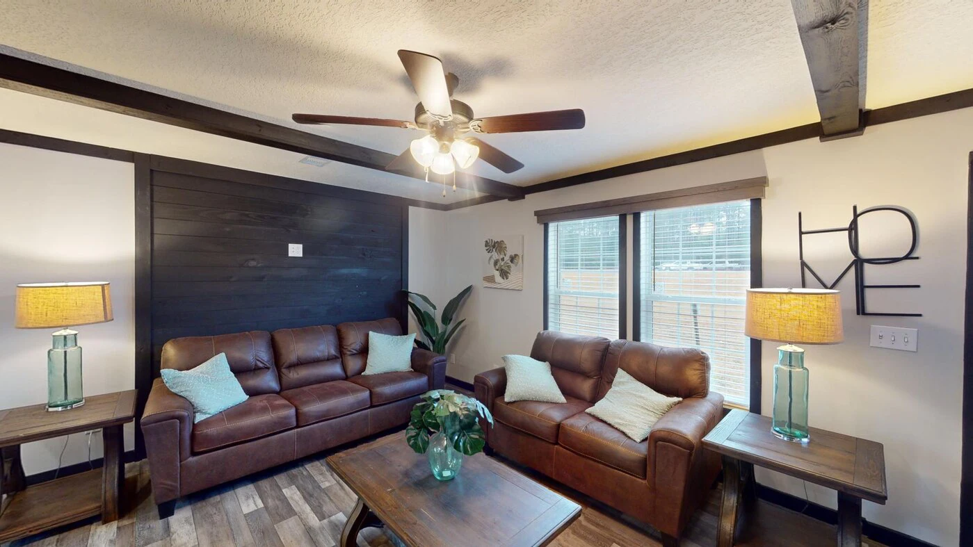 A living room with two brown leather sofas, a wooden coffee table, two side tables with lamps, a ceiling fan, and large windows with blinds.