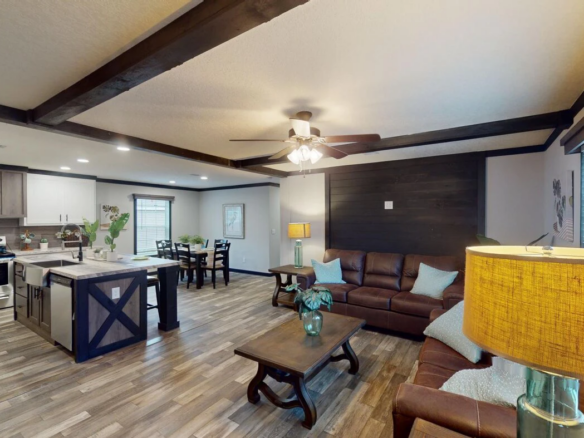 Open-concept living room and kitchen with wood beams, brown leather sofas, a central coffee table, an island, and modern appliances. Warm lighting and wood floors throughout.