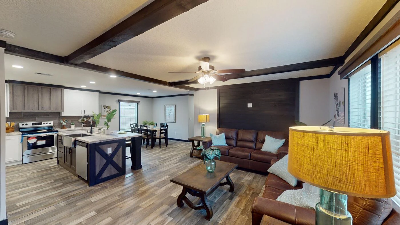 Open-concept living room and kitchen with wood beams, brown leather sofas, a central coffee table, an island, and modern appliances. Warm lighting and wood floors throughout.