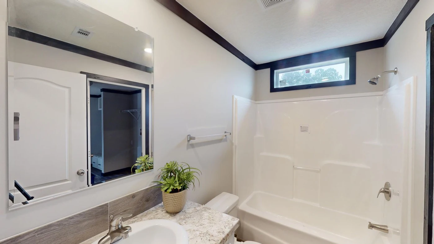 A modern bathroom with a bathtub-shower combo, a sink with a marble countertop, a wall mirror, a potted plant, and a small rectangular window above the shower.