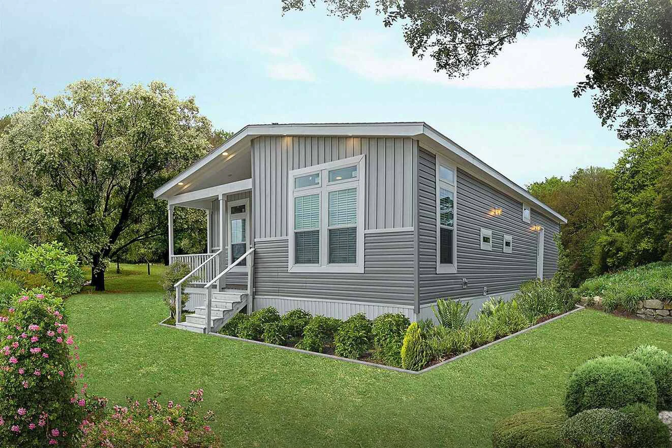 A gray single-story manufactured home with white trim, large windows, and a small front porch, surrounded by green grass, shrubs, and trees.