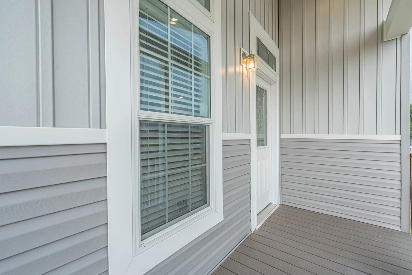 A close-up view of a house's exterior featuring gray siding, a white-framed window with blinds, a white door with glass, and a lit wall-mounted lantern.