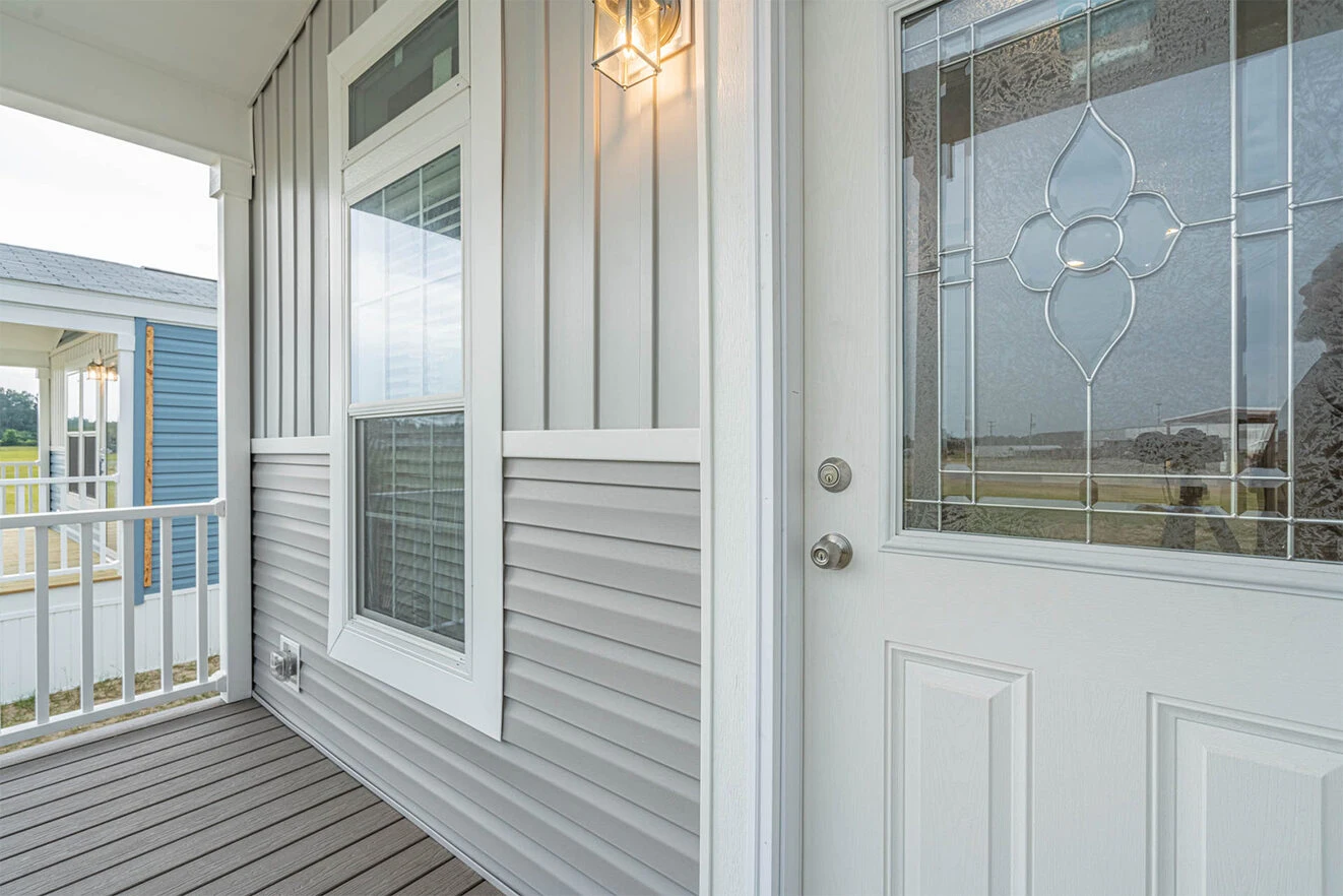 A close-up view of a house porch featuring gray siding, a window, a white front door with decorative glass, and a lit exterior light fixture.
